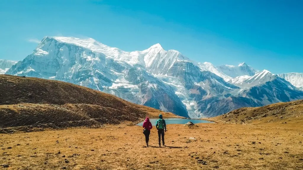 Una coppia che cammina verso il lago di ghiaccio, nell'ambito dell'Annapurna Circuit Trek, Himalaya, Nepal. La catena dell'Annapurna alle spalle, coperta di neve. Tempo sereno, erba secca, cime innevate. Alta quota